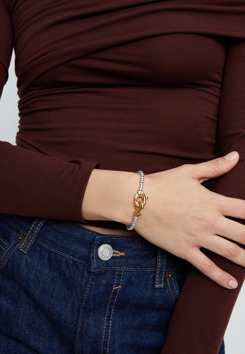 Pulsera de plata y oro con diseño de cadena entrelazada, cuentas metálicas y textura suave. Se lleva en una mano contra una blusa marrón.