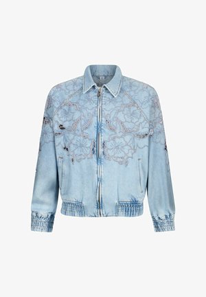 Veste en denim bleu clair avec fermeture éclair sur le devant, poignets et ourlet élastiques, présentant des motifs complexes de découpe en dentelle florale sur la poitrine et les manches.