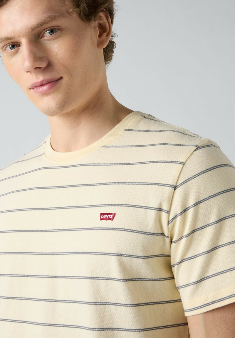Jeune homme portant un T-shirt crème à manches courtes avec de fines rayures horizontales grises et un petit logo Levi's rouge sur la poitrine.