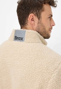 Giacca di pile beige con una finitura testurizzata, caratterizzata da un colletto alto e un'etichetta grigia sul retro che mostra "Brax" in lettere nere in grassetto.