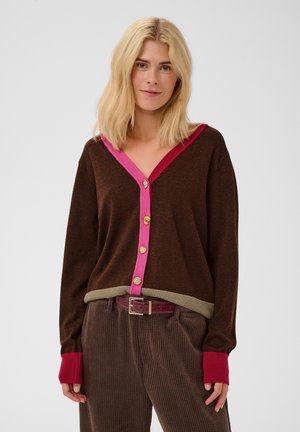 Frau mit blondem Haar, die eine braune Strickjacke mit rosa Besatz und einzigartigen Knöpfen trägt, kombiniert mit brauner Cordhose und einem bordeauxroten Gürtel.
