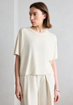 Jonge vrouw met kort bruin haar, gekleed in een loszittend, gebroken wit shirt met korte mouwen en bijpassende wijde broek, kijkend naar links tegen een effen achtergrond.