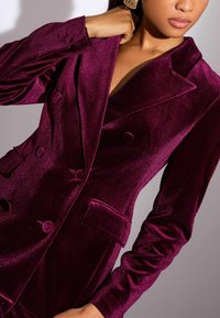 Plüss, mély burgundi blézer dupla soros gombos dizájnnal, nagy gombokkal, hajtókával és strukturált vállakkal. Csillogó textúra jellemzi.
