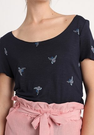 Femme portant un haut bleu marine à manches courtes avec de petits imprimés floraux et un pantalon taille haute rose noué avec une ceinture en tissu.