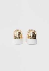 O pereche de adidași metalici aurii cu tălpi groase albe, cu logo-ul „GUESS” embosat pe toc, prezentată din spate pe un fundal simplu.