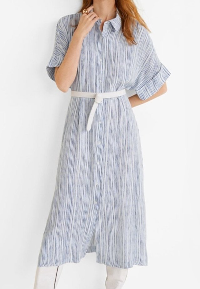 Femme portant une robe midi boutonnée rayée verticalement bleu et blanc, manches retroussées et ceinture blanche, debout sur un fond blanc.