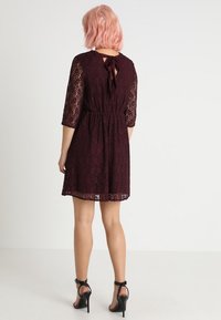 Robe en dentelle bordeaux avec des manches trois-quarts, taille élastique et un lien à l'arrière. Présente un ourlet au genou et une texture transparente.