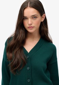 Donkergroene cardigan met een v-hals, geribbelde manchetten en vijf knopen aan de voorkant. Gebreide textuur, lange mouwen en een ontspannen pasvorm.