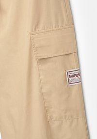 I pantaloni cargo beige presentano una tasca laterale e un'etichetta con la scritta "PACIFIC", con cuciture visibili e una superficie liscia e leggera.