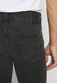 Kadar u krupnom planu osobe koja nosi crne traperice i bijelu košulju, prikazano je stražnje džep i označena kožna oznaka na pojasu.