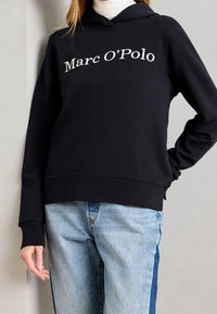 Czarna bluza z kapturem z białym logo "Marc O'Polo" umieszczonym na środku klatki piersiowej. Mieszanka bawełny, luźny krój, kieszeń kangurkowa, ściągacze przy mankietach.