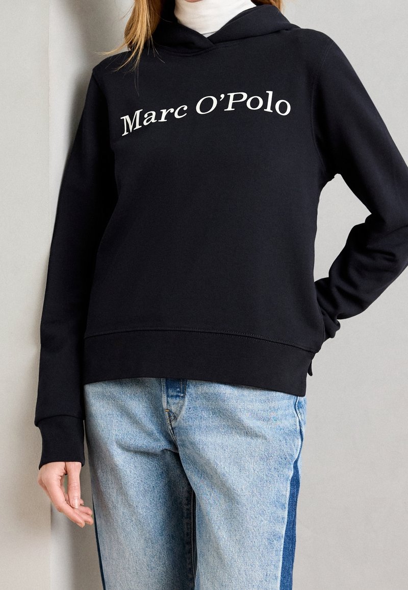 Czarna bluza z kapturem z białym logo "Marc O'Polo" umieszczonym na środku klatki piersiowej. Mieszanka bawełny, luźny krój, kieszeń kangurkowa, ściągacze przy mankietach.