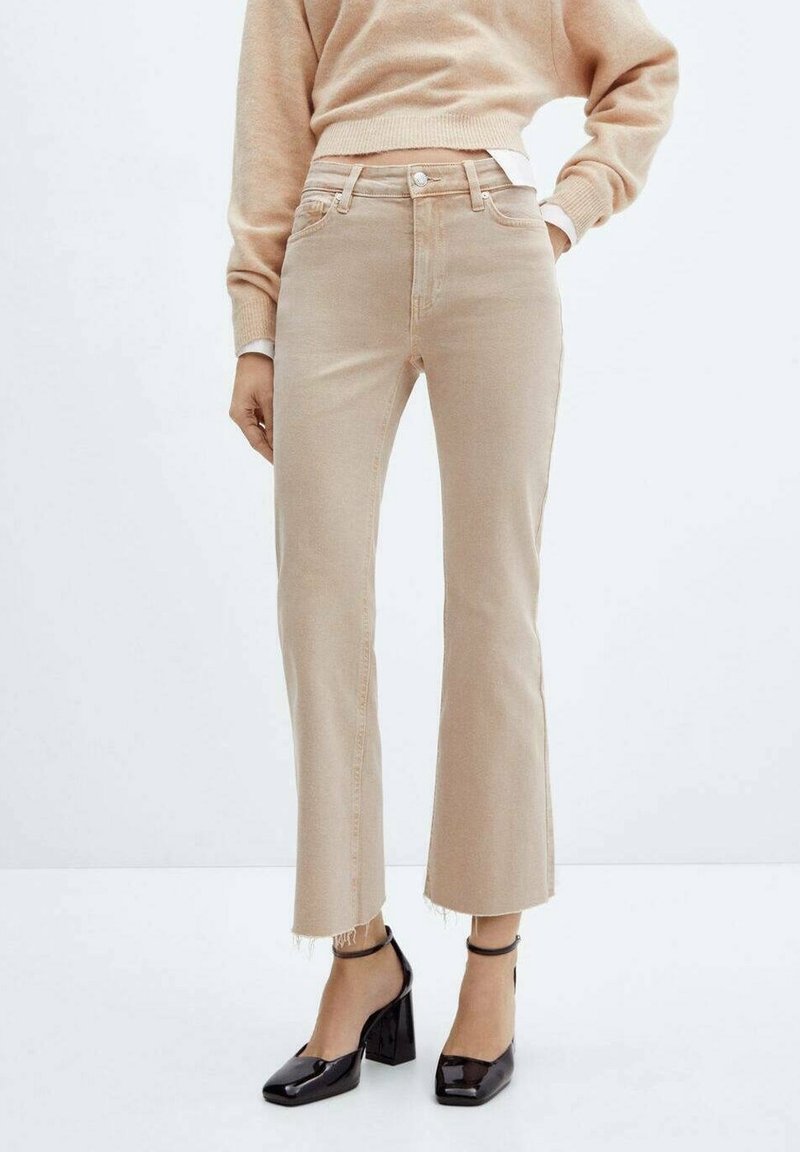 Jeans beige taille haute avec une jambe évasée, ourlet brut et fermeture à bouton. Portés avec des chaussures à talons blocs noires et un pull léger.