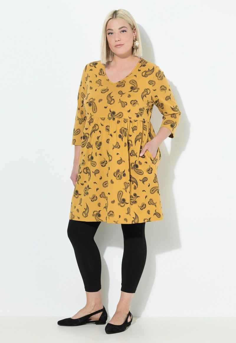 Donna in piedi con la mano in tasca indossa un vestito paisley giallo senape, leggings neri, scarpe nere e orecchini a forma di foglia dorata.