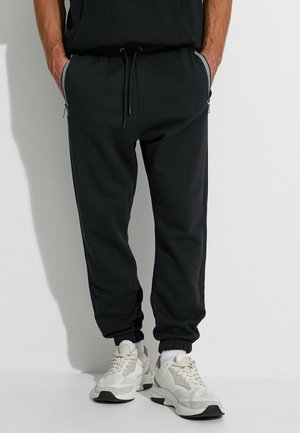 Schwarze Jogginghosen aus weichem Stoff, mit einem Kordelzug an der Taille, seitlichen Reißverschlusstaschen und gerippten Bündchen. Getragen mit hellen Sneakers.