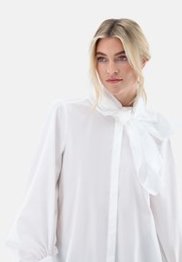 White blouse with long sleeves, soft fabric, bow tie detail at the neck, button closure, and gathered cuffs. Simple, elegant design.