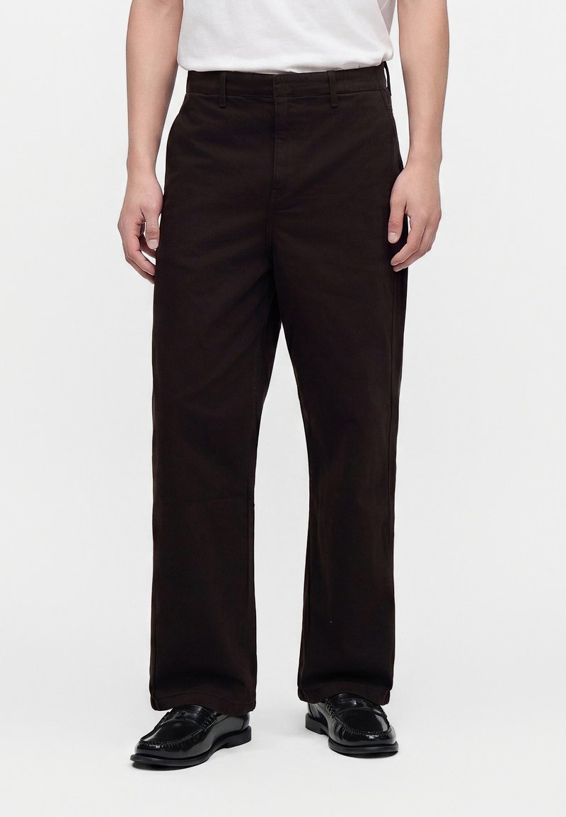 Homme portant un pantalon noir à jambe droite et des chaussures en cuir noir, debout devant un fond blanc uni.
