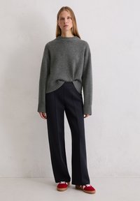 Szary wełniany sweter z prążkowanymi mankietami, zestawiony z granatowymi spodniami o szerokich nogawkach. Model nosi czerwone sneakersy z białymi akcentami, stojąc na jednolitym tle.