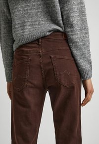 Jeans di denim marrone con due tasche posteriori, caratterizzati da un motivo a onde suttili nella cucitura. Abbinati a un maglione grigio lavorato a maglia.