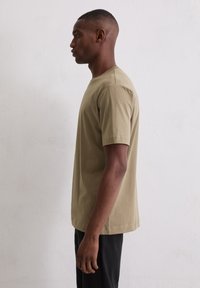 Man die in profiel staat, gekleed in een beige t-shirt met korte mouwen en zwarte broek, tegen een effen witte muur.