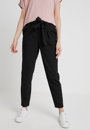 Kobieta ubrana w czarne spodnie w prążki z wiązanym paskiem w talii, różową koszulę z krótkim rękawem i białe trampki, stojąca na jednolitym tle.