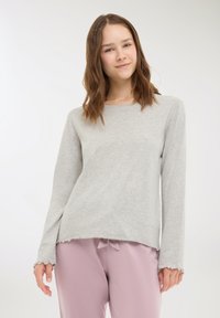 Maglia grigia chiaro a maniche lunghe realizzata in materiale morbido con scollatura arrotondata. Presenta polsini a coste e un orlo leggermente irregolare. Abbinata a pantaloni rosa.