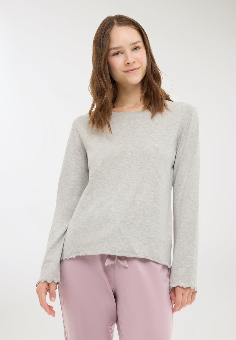 Maglia grigia chiaro a maniche lunghe realizzata in materiale morbido con scollatura arrotondata. Presenta polsini a coste e un orlo leggermente irregolare. Abbinata a pantaloni rosa.