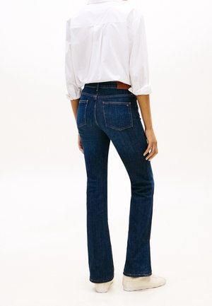 Personne portant un jean bootcut bleu foncé, une chemise blanche à manches longues retroussées, et des baskets blanches, debout, de dos.