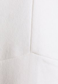 Bílá mikina s texturovaným vzorem, která má překrývající se části látky a žebrovaný lem. Hladký povrch a jemný vzhled.