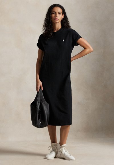 Μαύρο βαμβακερό φόρεμα T-shirt με κοντά μανίκια που είναι γυρισμένα, ίσια κοπή, μήκος μέχρι το γόνατο, με μικρό λευκό λογότυπο, συνδυασμένο με λευκά ψηλά αθλητικά παπούτσια και μαύρη τσάντα.