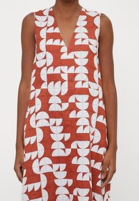 Ärmlös klänning i rostfärg med vitt geometriskt mönster. Har en V-ringning och en lös form. Tyget verkar lätt och texturerat.