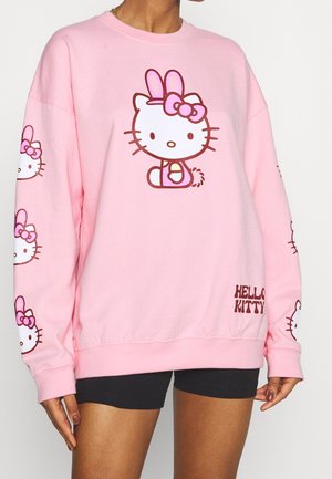 Mujer con sudadera rosa que tiene un gran personaje de Hello Kitty con orejas de conejo y caras más pequeñas de Hello Kitty en las mangas, combinada con pantalones cortos negros.