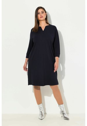 Dunkelblaues knielanges Kleid mit Dreiviertelärmeln, das einen V-Ausschnitt aufweist und eine glänzende Textur hat; kombiniert mit glänzenden silbernen Stiefeletten.
