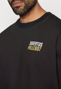 Sudadera negra con cuello redondo, que presenta un logo en blanco y amarillo "JUVENTUS MILLE897" en el pecho. Textura de tela suave.