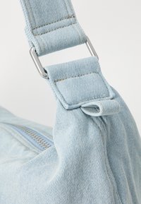 Modra denim ramenska trak s srebrnim okovjem, ki vključuje teksturirano tkanino in vidne šive, pritrjena na rob mehke torbe.