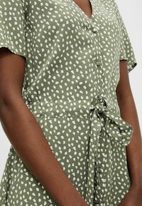 Robe verte avec un motif à pois blancs, à manches courtes, décolleté en V, détails de boutons et taille nouée, fabriquée dans un textile doux.