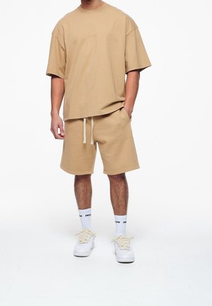 T-shirt oversize beige con scollatura rotonda, abbinata a pantaloni corti beige coordinati. Sneakers bianche e calze con testo nero completano l'outfit.