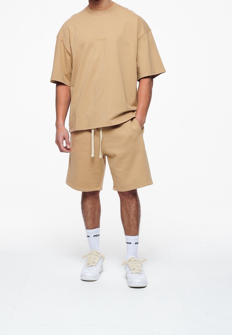 Beige oversized t-shirt med rund halsudskæring, parret med matchende beige shorts. Hvide sneakers og strømper med sort tekst fuldender outfittet.