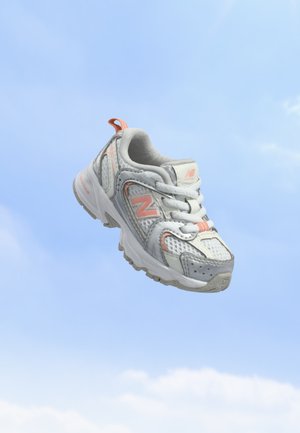 Άσπρο και γκρι παπούτσι για τρέξιμο New Balance με ροδακινί λεπτομέρειες που αιωρείται πάνω από έναν καθαρό γαλανό ουρανό με ελαφριές νεφώσεις.