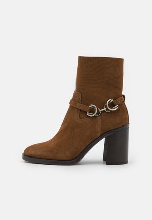 Botte cheville en daim brun avec un tige en tricot côtelé, un bout carré et un talon épais. Ce modèle présente une sangle décorative avec des anneaux en métal argenté.