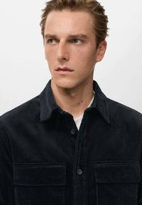 Camicia in velluto a coste nera con colletto, chiusura frontale con bottoni e due tasche sul petto. Presenta una texture a coste verticali e una finitura liscia.