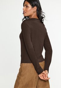 Haut marron à manches longues avec un design côtelé ajusté. Encolure ronde et épaules sans coutures. Associé à une jupe en daim brun clair.