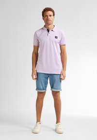 Světle fialová polo tričko s černým límcem a logem na náprsní kapse, doplněné o ohrnuté džíny šortky a bílé tenisky. Jednoduchý, neformální design.