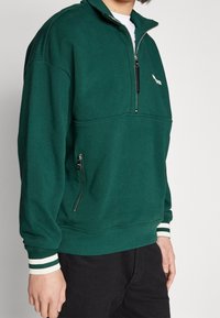 Sudadera verde con cremallera hecha de una tela suave, con medio cuello, puños a rayas blancas y verdes, y un bolsillo lateral con cremallera.