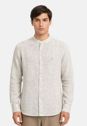 Giovane uomo con una camicia beige chiaro a maniche lunghe con collo alla coreana, in piedi davanti a uno sfondo neutro.
