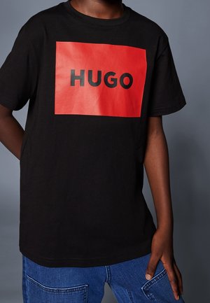 Persona que lleva una camiseta negra con un gran rectángulo rojo y la palabra "HUGO" en letras negras en el pecho, combinada con vaqueros azules.