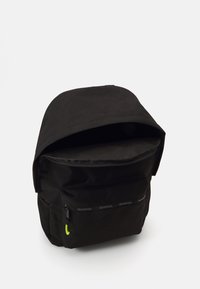 Mochila negra hecha de tela resistente, con un bolsillo frontal con cremallera y un logotipo reflectante de Reebok en la parte delantera. Incluye tirantes ajustables.
