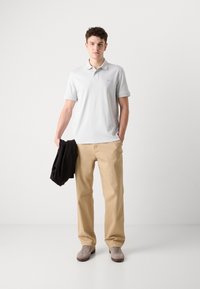 Camisa polo cinza claro, calças bege, sapatos cinzentos, segurando uma jaqueta preta. Design simples, mangas curtas, tecido suave, marcação mínima.