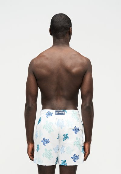 Άνδρας με σκουρόχρωμο δέρμα, με μυώδη πλάτη, φοράει λευκά μαγιό με μπλε σχέδια χελωνών, στέκεται μπροστά σε ένα απλό φωτεινό φόντο.