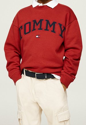 Person iført en rød sweater med teksten "TOMMY" og et lille flaglogo, hvid skjorte med krave under, sort bælte og lyse beige cargobukser.